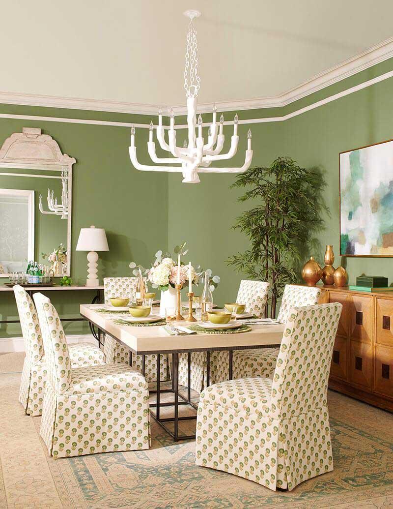 A lifestyle photo of a pale wood with a metal base dining table with six patterned slip covered dining chairs around it. A white chandelier and a green wall.